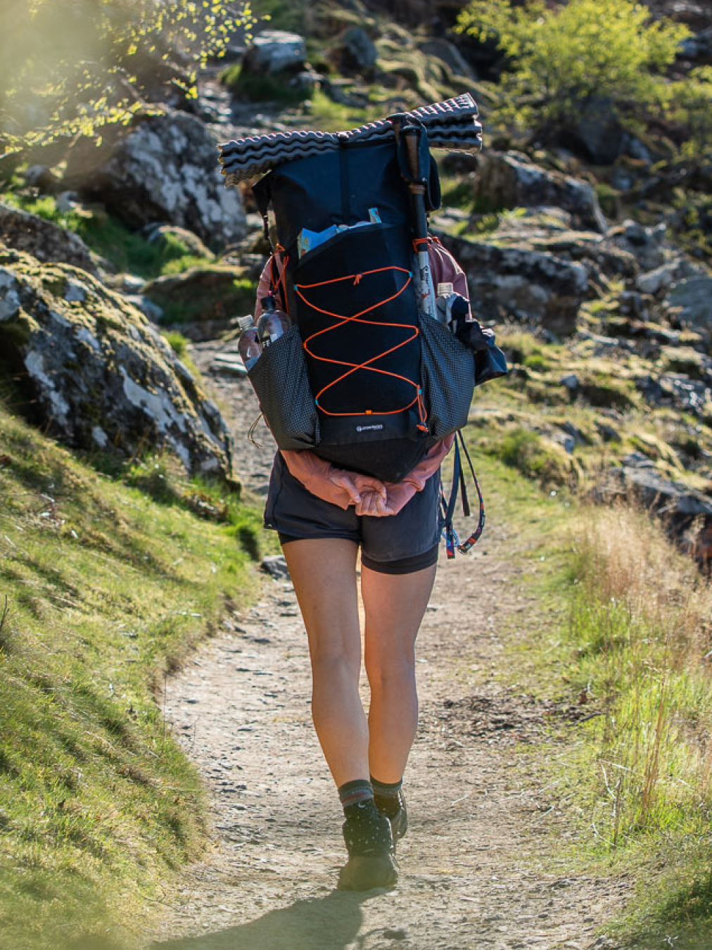 A hiker viewed from behind on a rocky dirt trail, wearing a fully loaded Prospector.