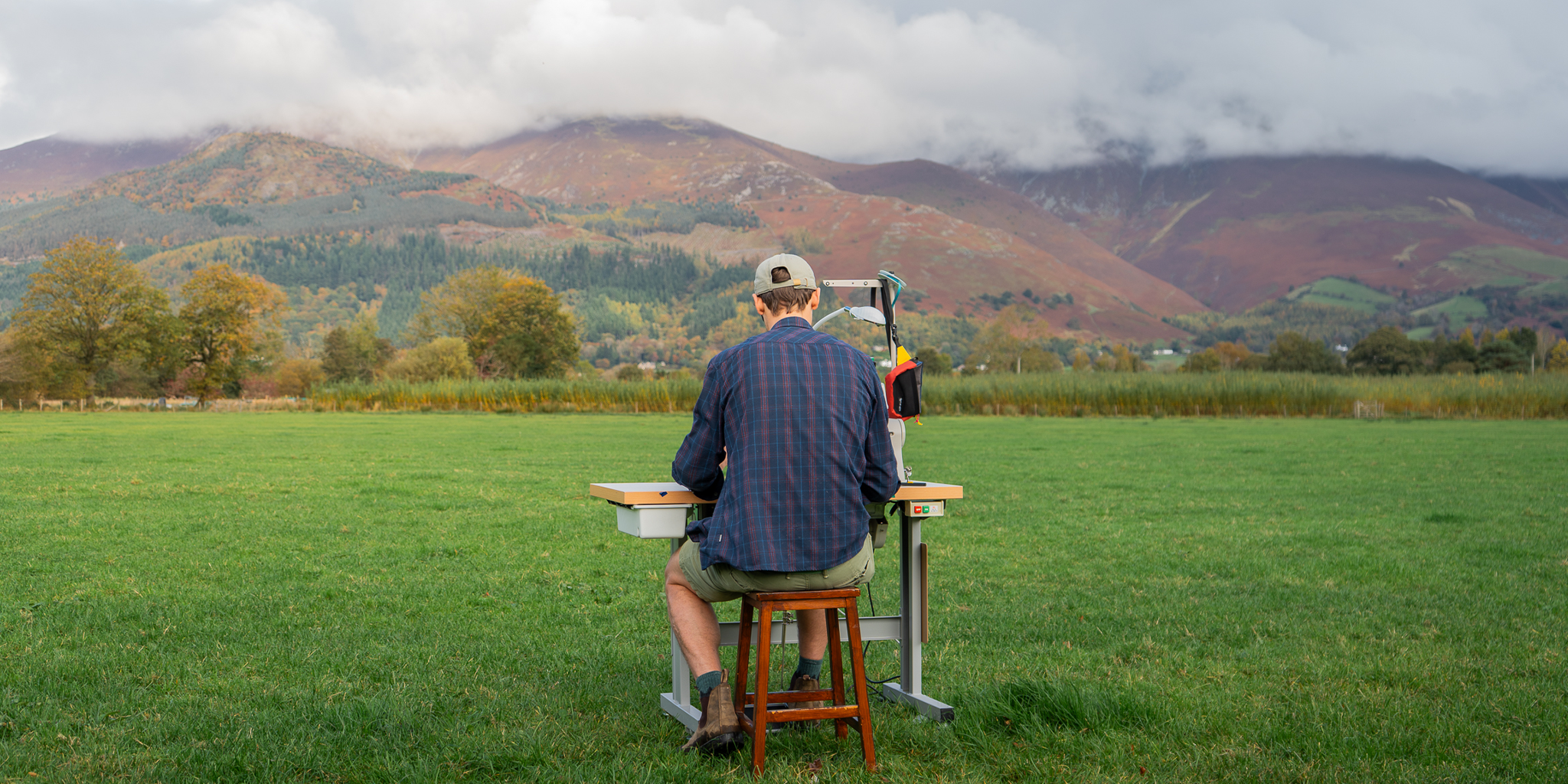 Tom, Atom Packs founder, sitting at a sewing table in a field with mountains in the background